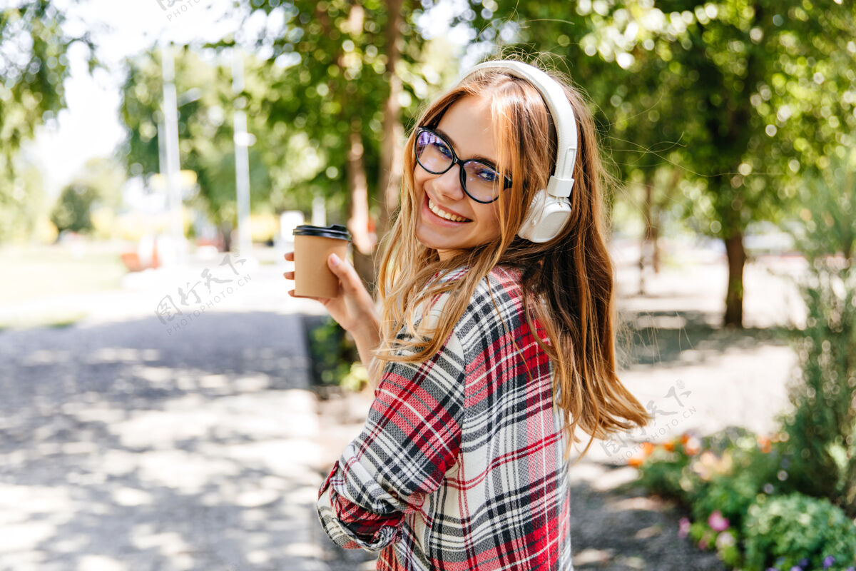 休息在公园里喝咖啡的幽默女孩戴着眼镜的金发女人在听大自然的音乐放松阳光公园