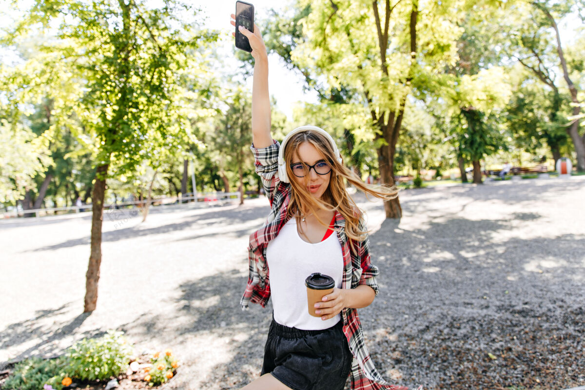 青少年悠闲的女人端着一杯咖啡在公园里翩翩起舞温文尔雅的金发女郎戴着眼镜 在早晨欣赏大自然的音乐人欢笑休闲