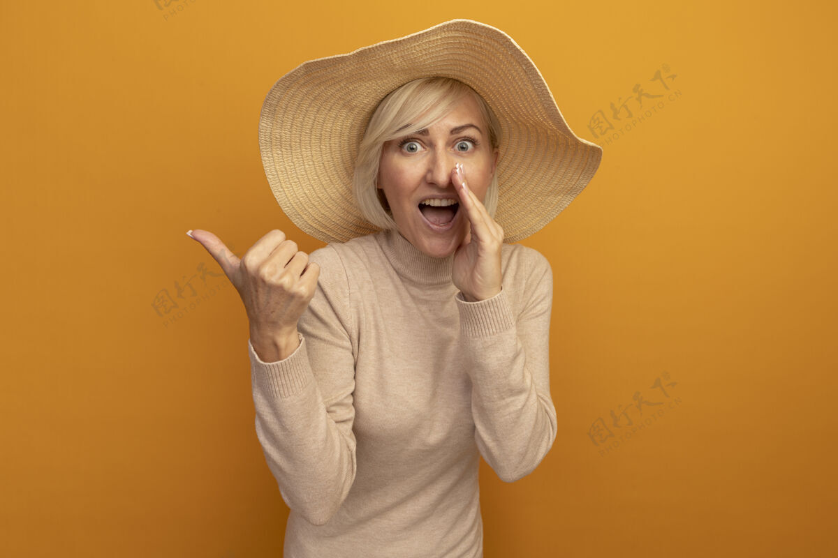 海滩兴奋的金发斯拉夫女人 戴着沙滩帽 手紧握着嘴巴 指着橙色的边漂亮金发女人