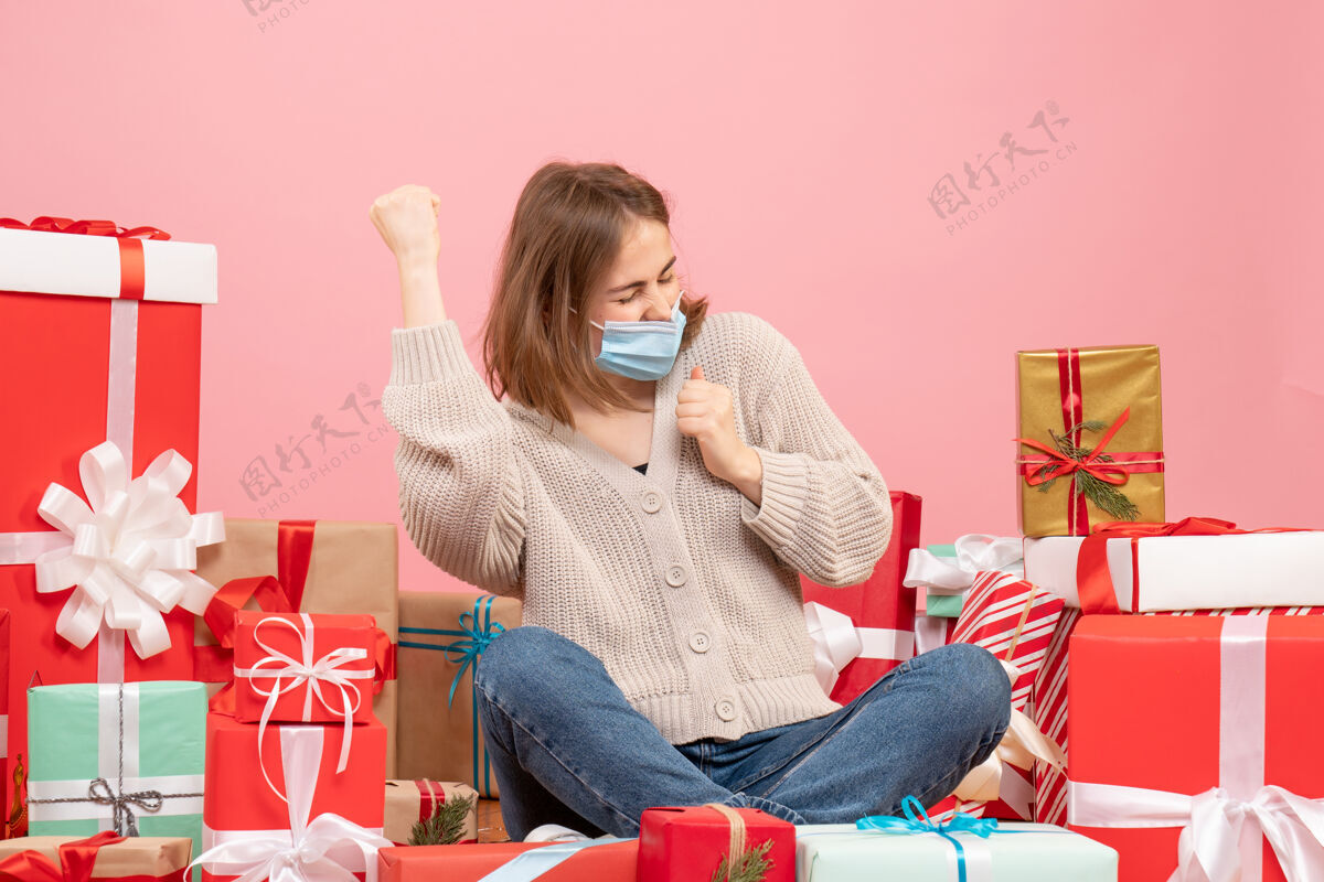 年轻女性正面图：年轻女性戴着面具围坐在圣诞礼物旁边礼物坐着病毒