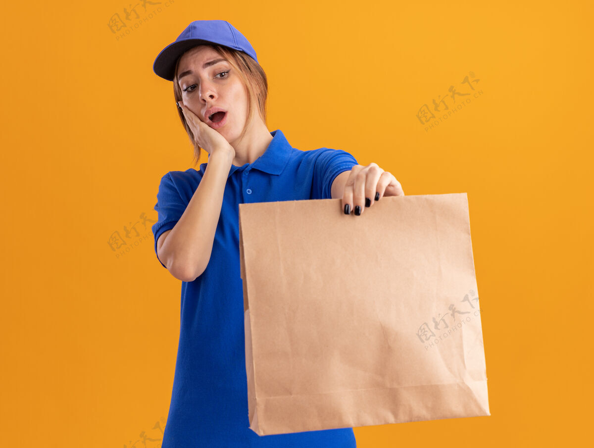 制服焦急的年轻漂亮的女送货员 穿着制服 把手放在脸上 拿着桔黄色的纸包看着姿势手包装