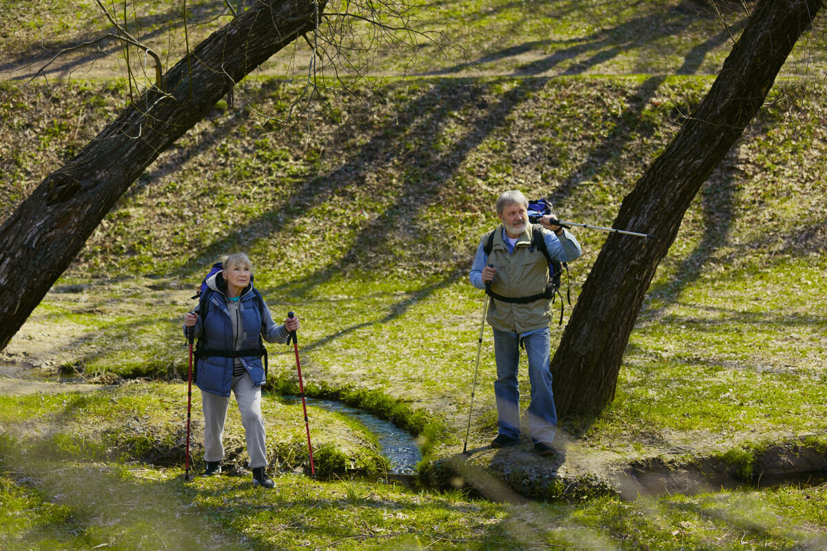 成熟我们可以一起做一对穿着旅游服装的老年夫妇在阳光明媚的日子里走在小溪边的绿色草坪上旅游的理念 健康的生活方式 放松和团聚田园诗女性男性