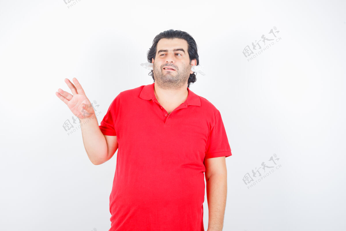 男人帅哥举起手 眨着眼睛 站着 耳朵里塞着餐巾 穿着红色t恤 看上去优柔寡断前视图不确定沉思头