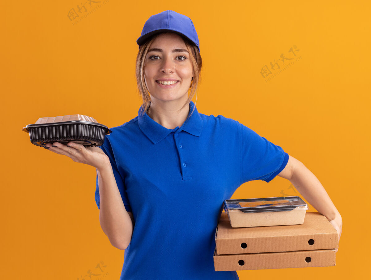 姿势微笑着的年轻漂亮的送货女孩穿着制服拿着纸食品包装和容器放在橙色的比萨饼盒上制服比萨饼容器
