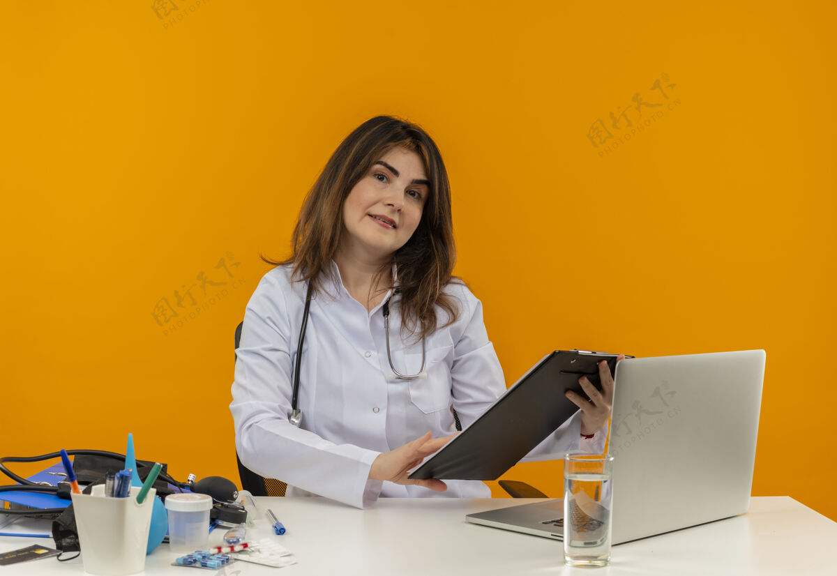 成人高兴的中年女医生穿着医用长袍和听诊器坐在办公桌旁 拿着医疗工具的剪贴板和手提电脑拿着剪贴板 看上去与世隔绝人橙色拿着