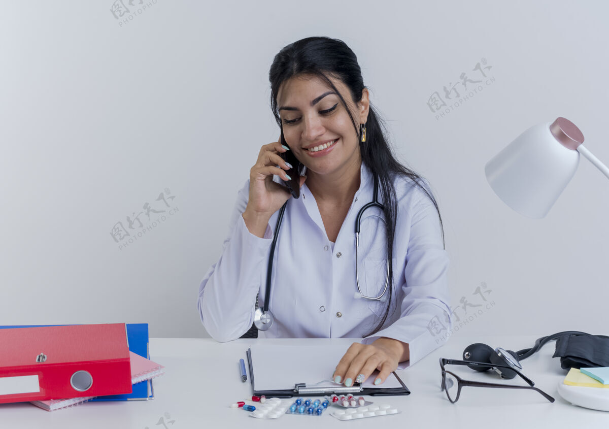 下来面带微笑的年轻女医生穿着医用长袍和听诊器坐在办公桌旁 手里拿着医疗工具 手放在剪贴板上 一边打电话一边往下看女性人桌子