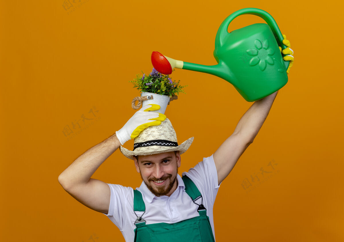 人身着制服的年轻英俊的斯拉夫园丁面带微笑 戴着帽子和园艺手套 头上举着花盆 用水壶浇水 看上去很孤独帽子橙色英俊