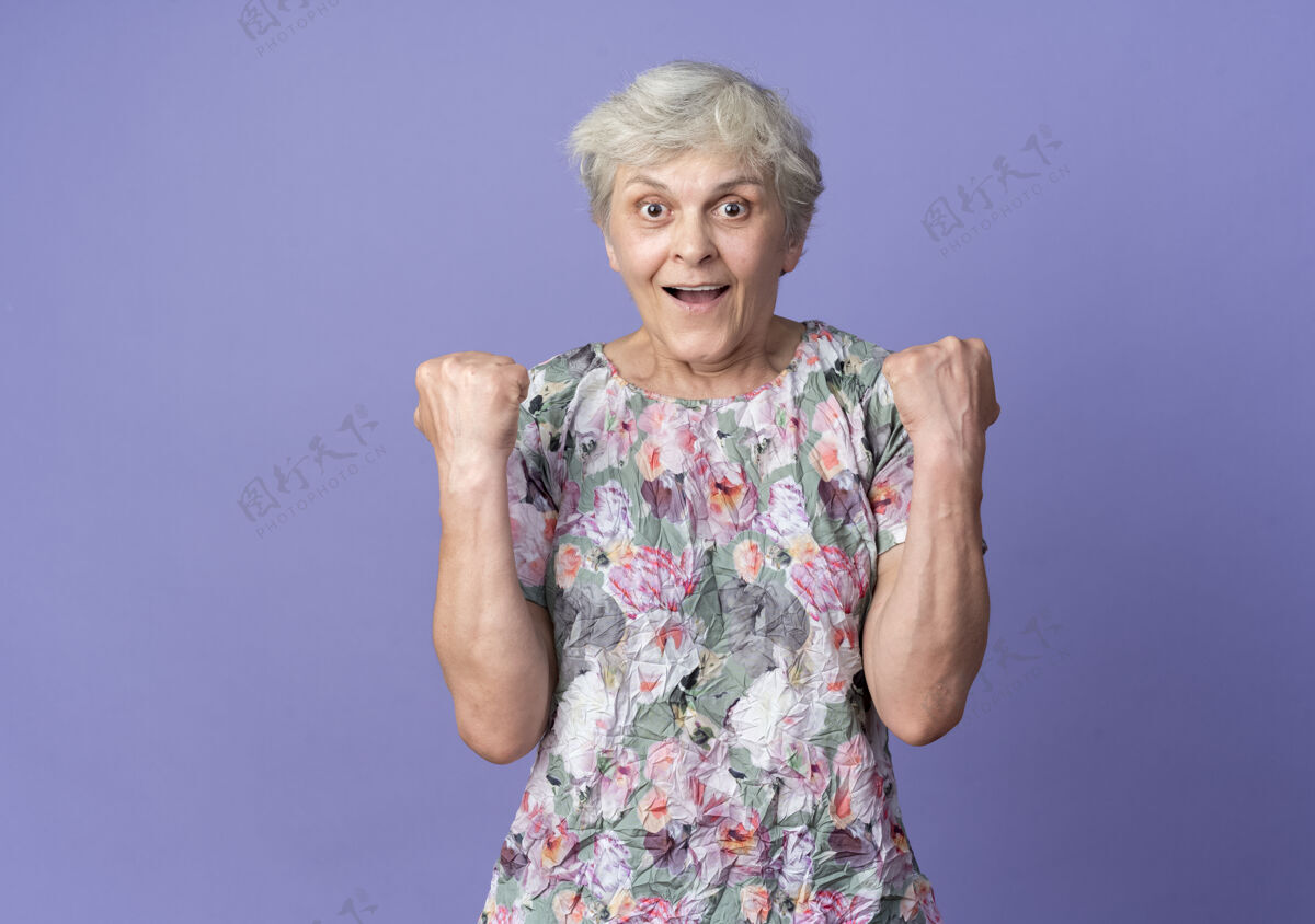 老兴奋的老妇人在紫色的墙壁上保持拳头看起来孤立老拳头兴奋
