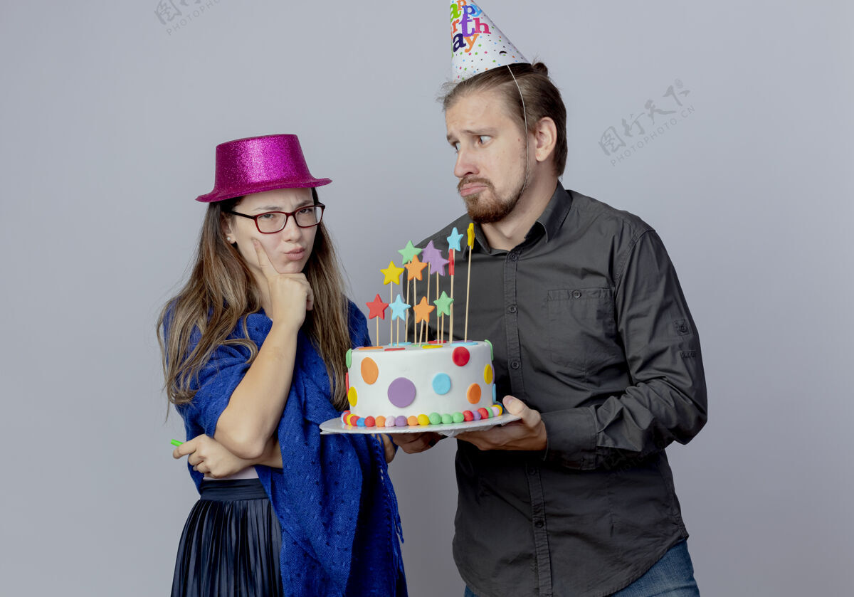 蛋糕戴着生日帽的焦急帅哥手里拿着蛋糕 看着恼怒的戴着粉红色帽子的戴着眼镜的年轻女孩 手里拿着口哨 手放在隔离在白墙上的下巴上眼镜帽子穿