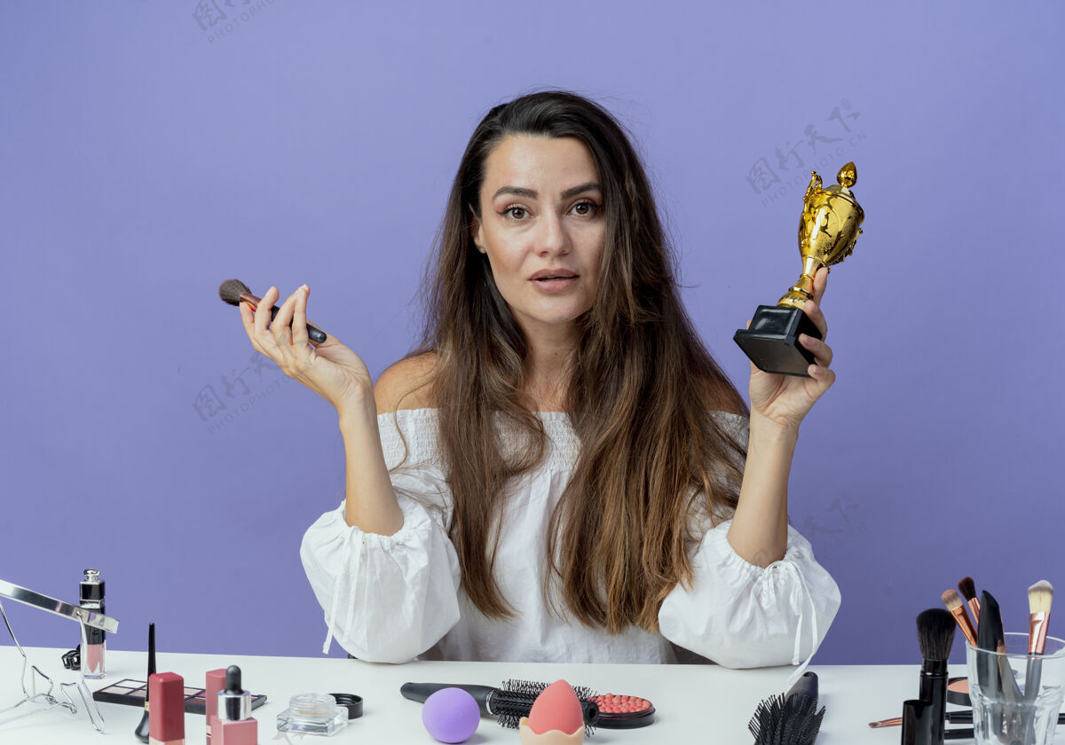 坐着自信美丽的女孩坐在桌子旁 拿着化妆工具 把化妆刷和优胜者杯孤立地放在紫色的墙上美丽工具桌子