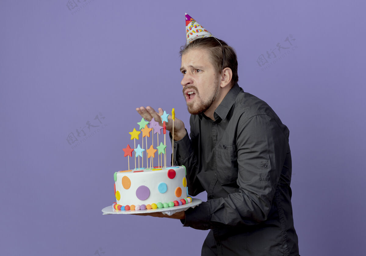 男人恼怒的帅哥戴着生日帽站在一边 拿着蛋糕指着紫色的墙上 孤零零的站着指着帽子