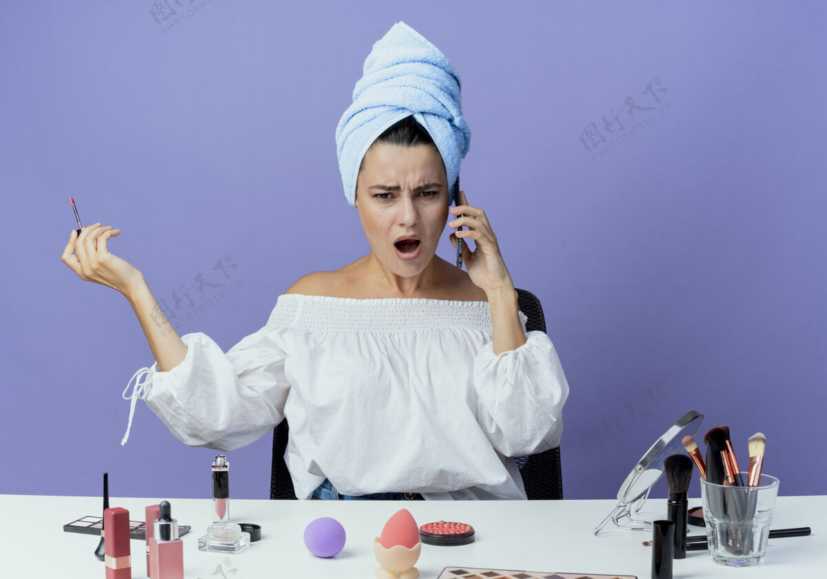 美丽恼人的漂亮女孩裹着发巾坐在桌子上用化妆工具夹着唇彩在电话里对着紫墙隔绝电话女人烦恼包裹