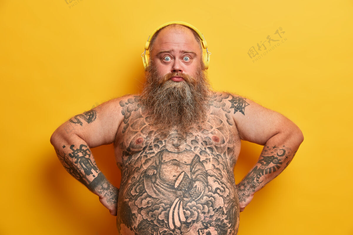 体重照片中的愁眉苦脸 满脸胡须的男人表情惊讶地戴着耳机听着最喜欢的歌 双手放在臀部 肚子大得发胖 身上有纹身 隔离在黄色的墙上大大情绪