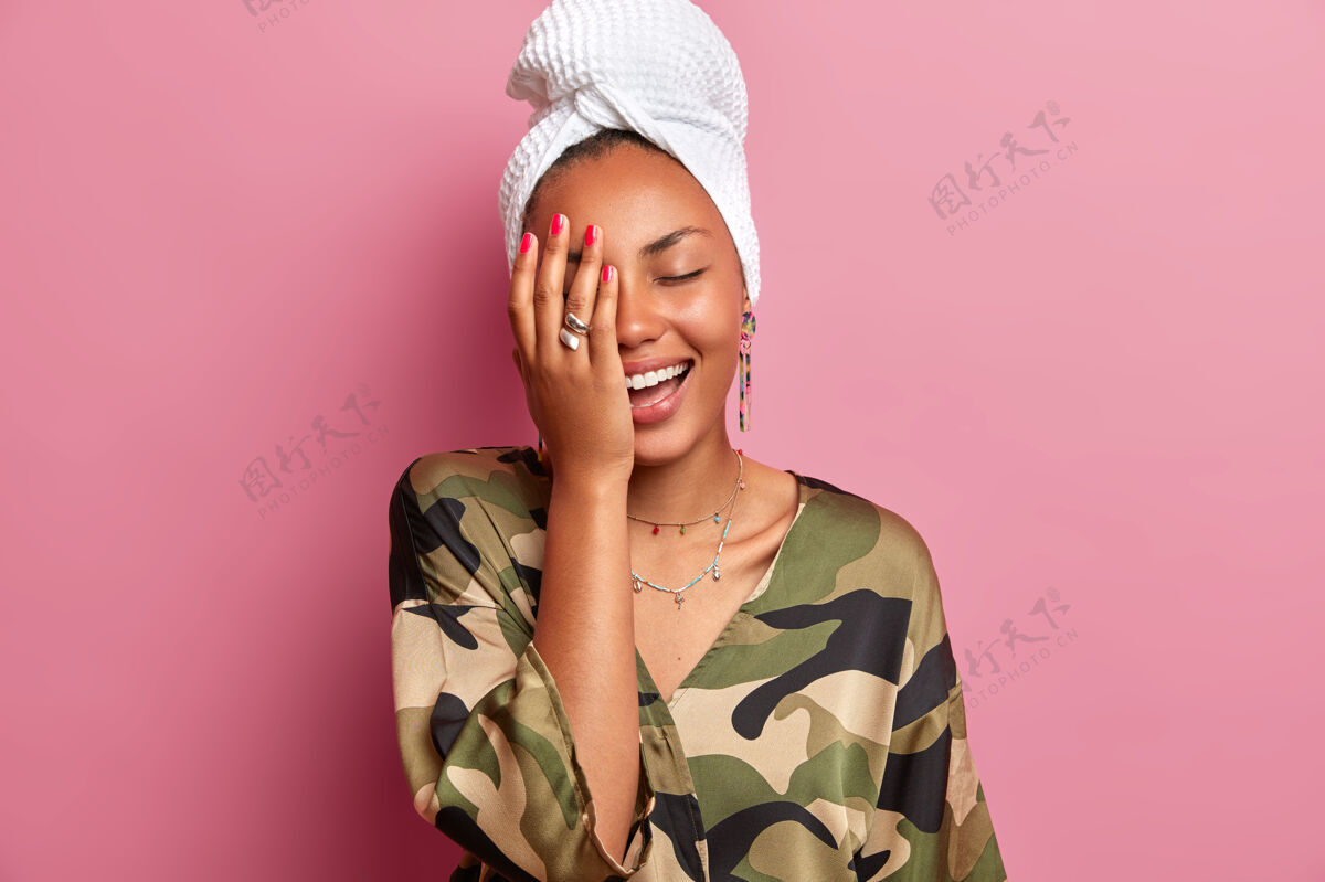 风格家庭式概念快乐的年轻女子积极地咯咯笑 使脸掌心 有健康干净的皮肤后 淋浴和美容程序 穿着长袍 姿势对粉红色的墙壁快乐水疗美容