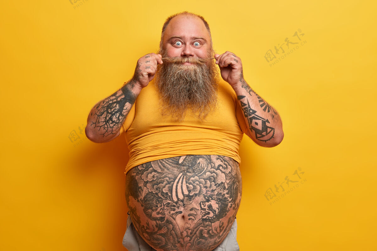腹部横拍搞笑的胖家伙 大肚子 手臂和腹部纹身 扭胡子 穿着黄色t恤 肥胖 因为喝了很多啤酒 吃垃圾食品胖懒男年轻体重阳刚