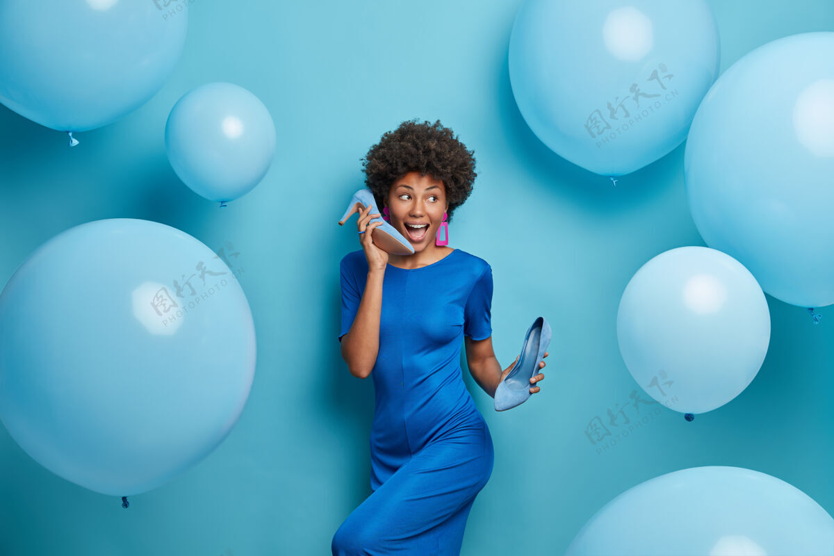 气球充满灵感的开朗女性愉快地无忧无虑地跳舞 有派对气氛 享受音乐 身着蓝色鸡尾酒礼服 手拿鞋 在气球的包围下度过自由的庆祝时光黑发享受时尚