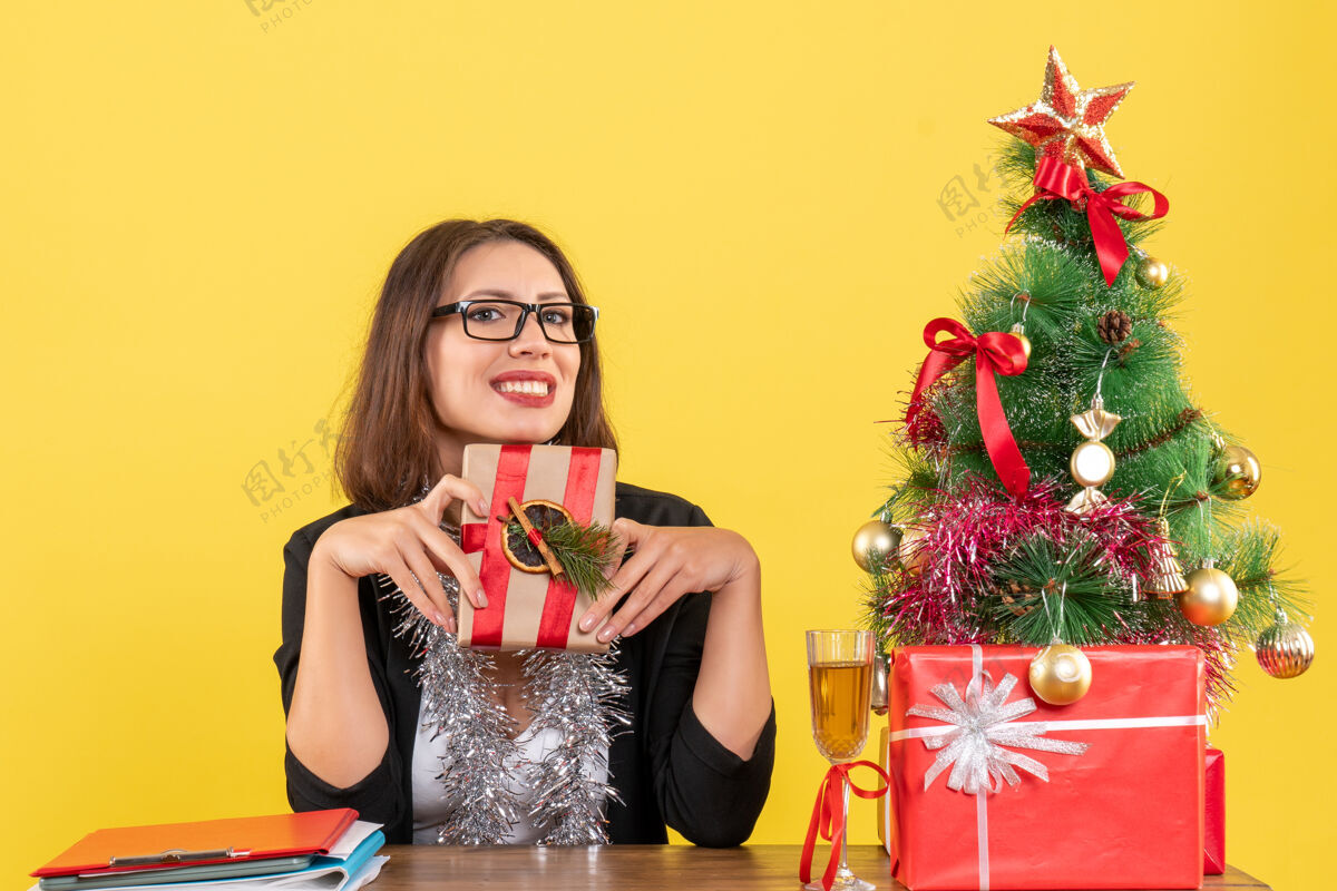 树一位身着西装 戴着眼镜 面带微笑的商务女士手里拿着礼物 坐在一张桌子旁 桌子上放着一棵圣诞树脸坐模型