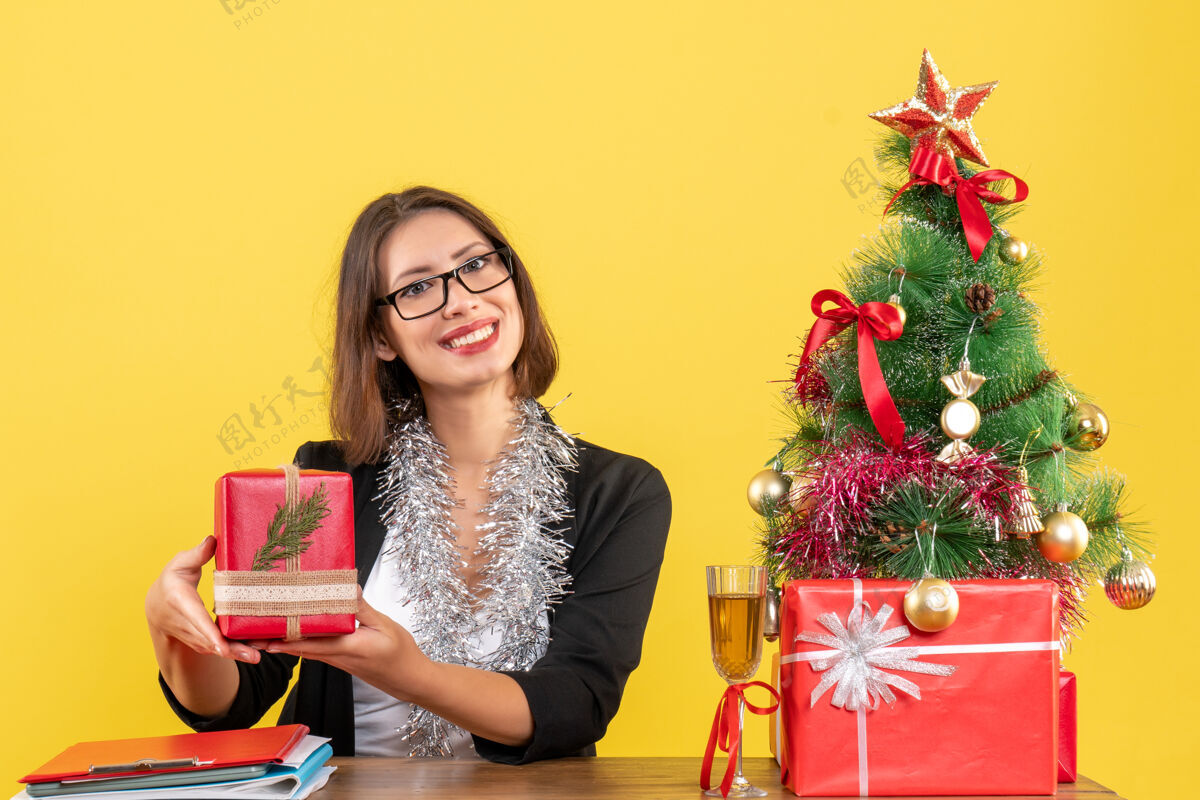 礼物一位身着西装 面带微笑的商务女士戴着一副眼镜 展示着她的礼物 坐在一张桌子旁 桌子上放着一棵圣诞树快乐桌子人