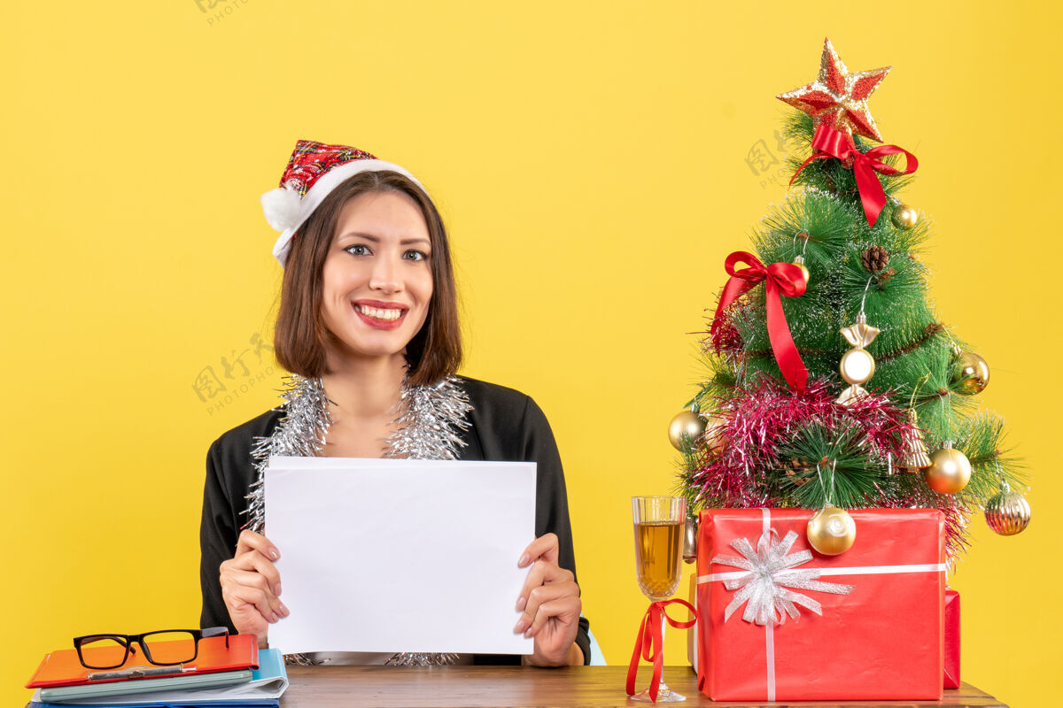 工作一位身着西装 戴着圣诞老人帽 戴着新年装饰品 面带微笑的商务女士独自一人拿着文件 坐在一张桌子旁 桌子上放着圣诞树抱着西装笔记本电脑
