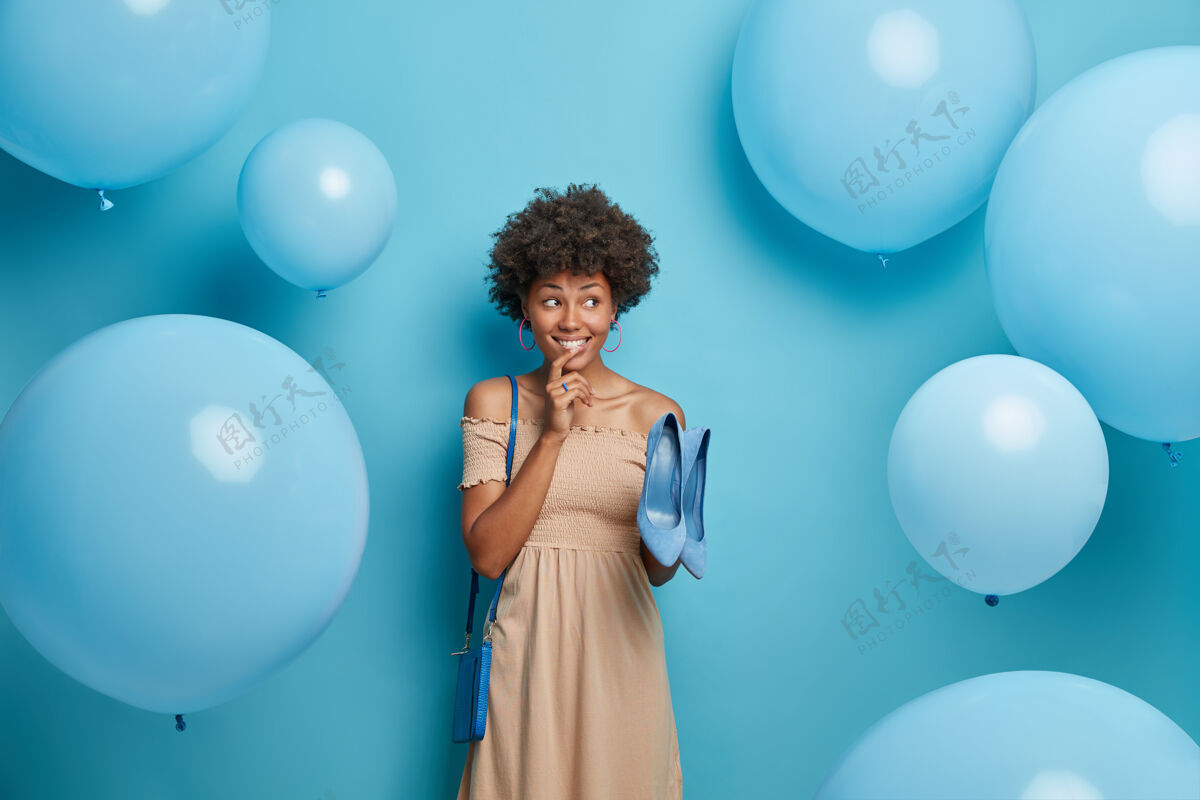 卷发正黑皮肤的女人穿着优雅的长裙 选择蓝色的鞋子来装包 在社团聚会上打扮得神采奕奕 靠着蓝色的墙壁 到处飞着气球时尚充气肖像