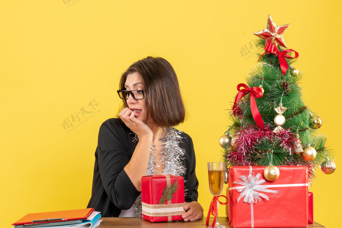 微笑好奇的商务女士穿着西装 戴着眼镜 手里拿着礼物 低头坐在一张桌子旁 桌子上放着一棵圣诞树快乐好奇坐着