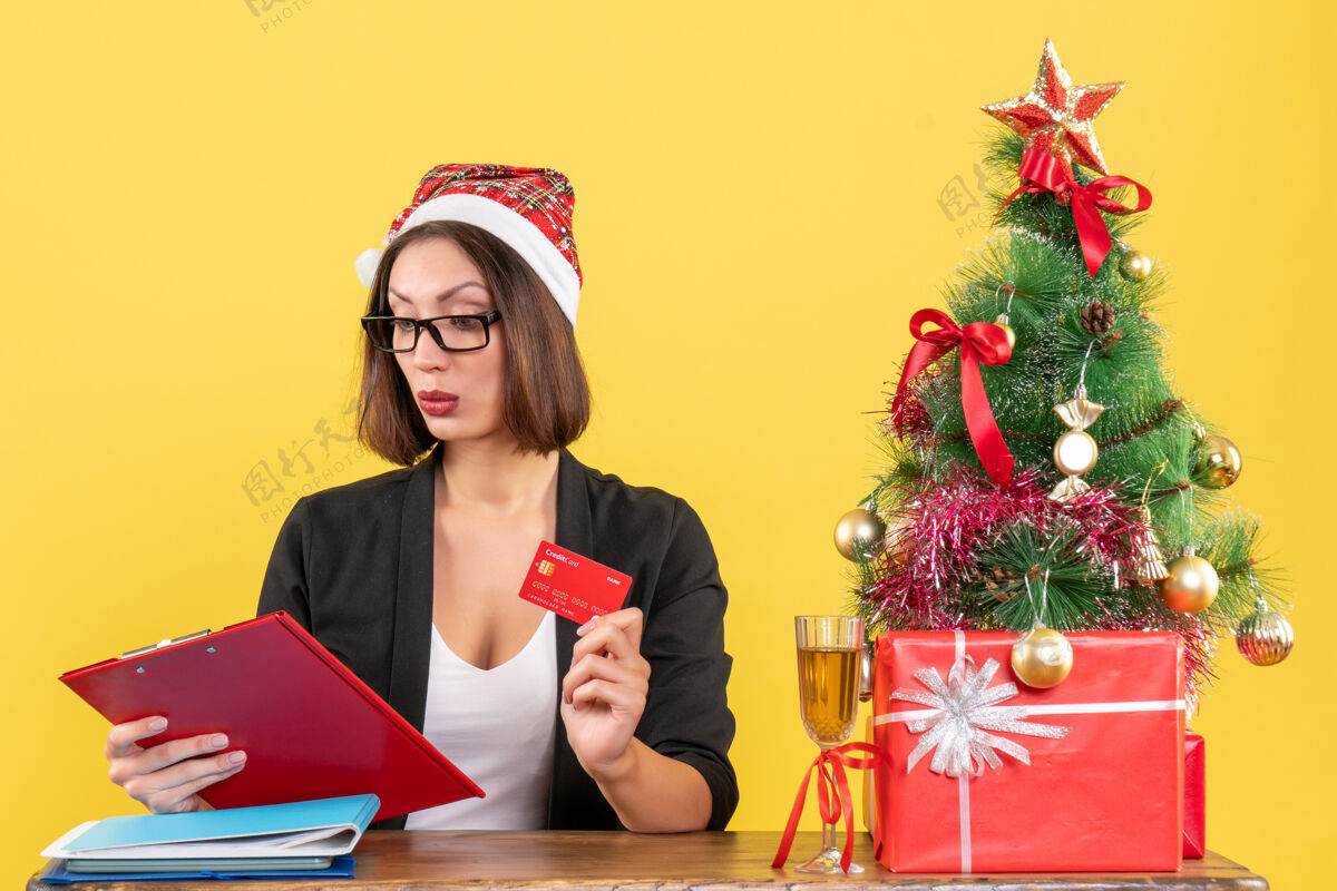 人集中迷人的女士穿着西装 戴着圣诞老人的帽子和眼镜 在黄色的办公室里出示银行卡圣诞老人快乐人