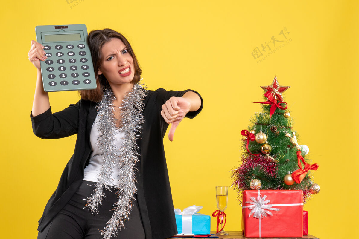 快乐圣诞气氛紧张美丽的女士站在办公室里 拿着计算器在办公室里做出消极的黄色手势晚餐礼服肖像时尚