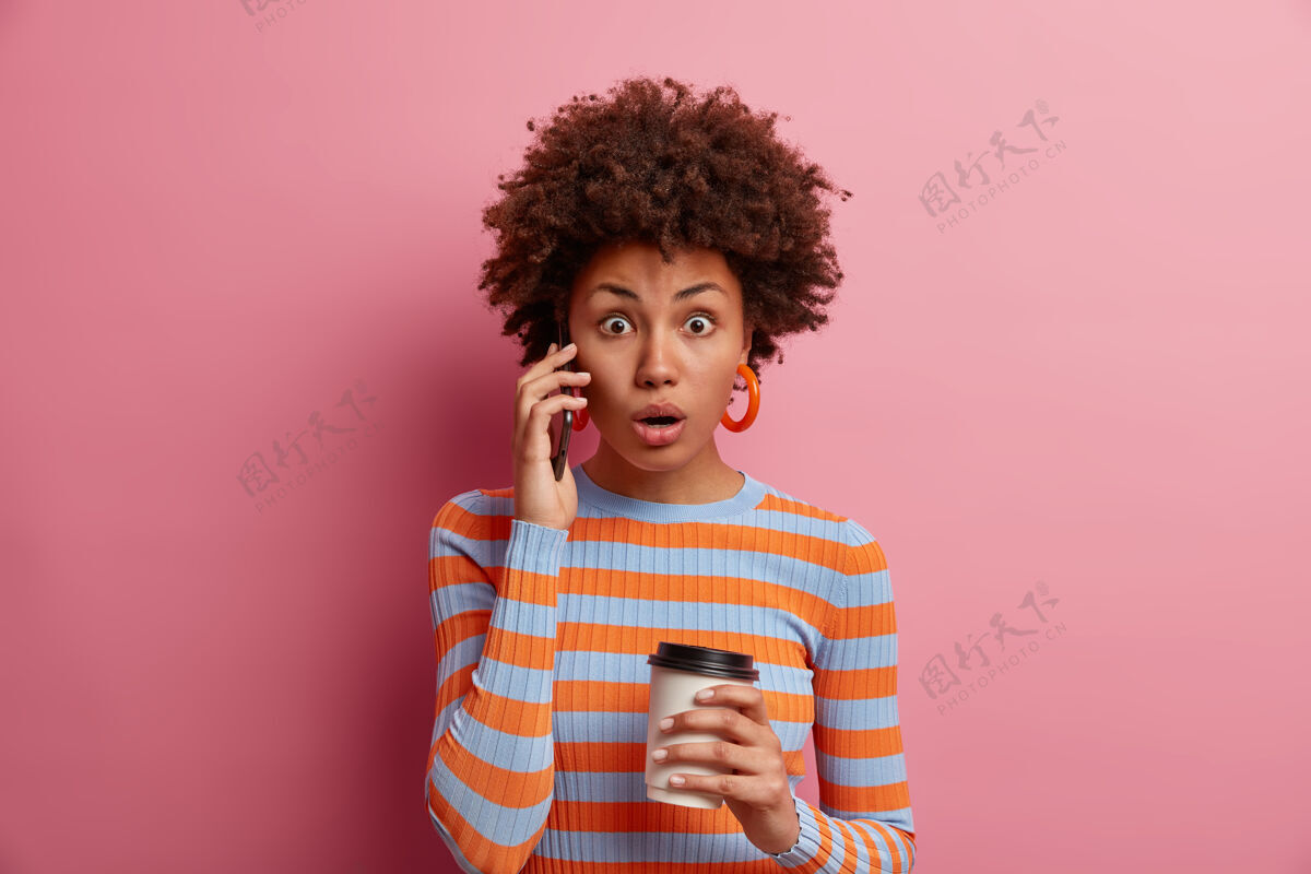 新闻震惊的女人听到新鲜的好消息 通过电话交谈 惊讶地张嘴 拿着外卖咖啡 表示惊讶 穿着休闲条纹套头衫 孤立在粉红色的墙上说话肖像套装