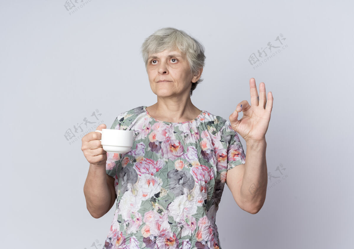 手势自信的老妇人拿着杯子 在白色的墙上做手势“ok”手势女人自信老人