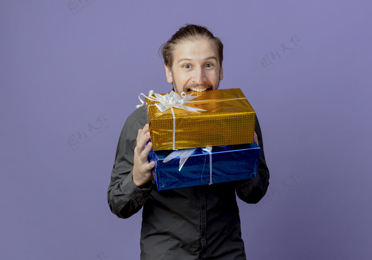 咬快乐的帅哥拿着和咬着紫色墙上看起来孤立的礼盒礼物帅气紫色