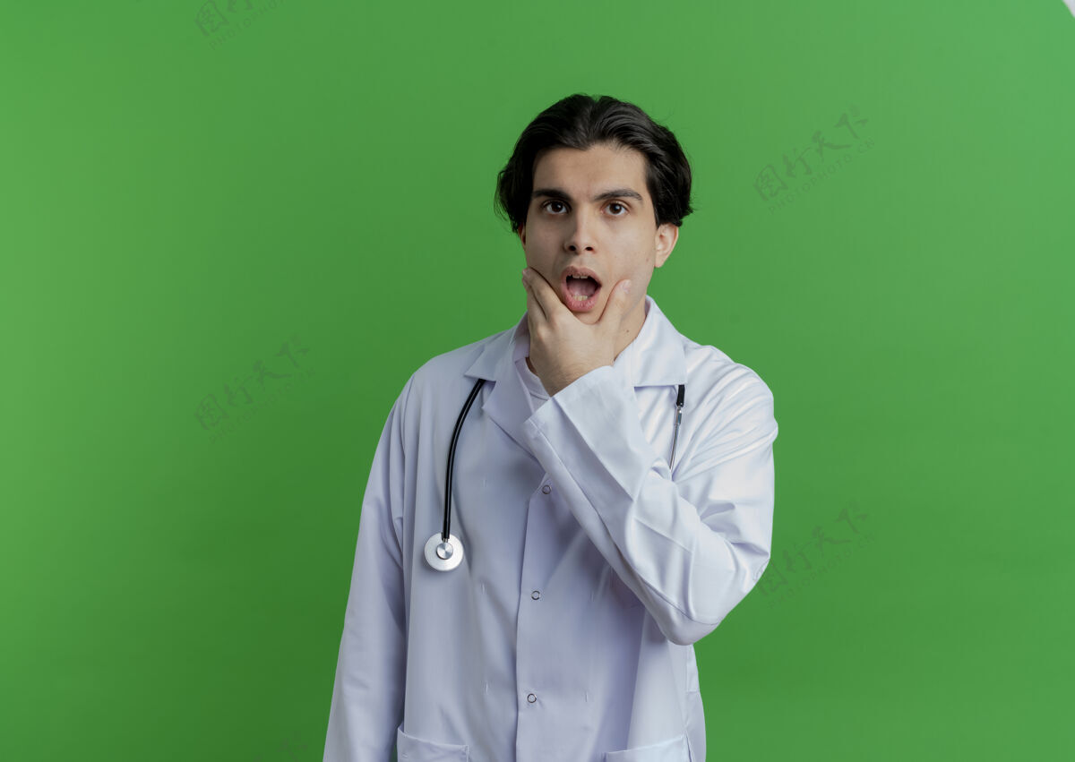 穿惊讶的年轻男医生穿着医用长袍 手持听诊器托着下巴人男人年轻