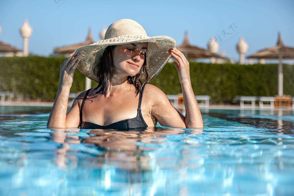 乐趣一个戴草帽的快乐女孩在阳光明媚的天气里沐浴在泳池里的特写镜头度假游泳肖像