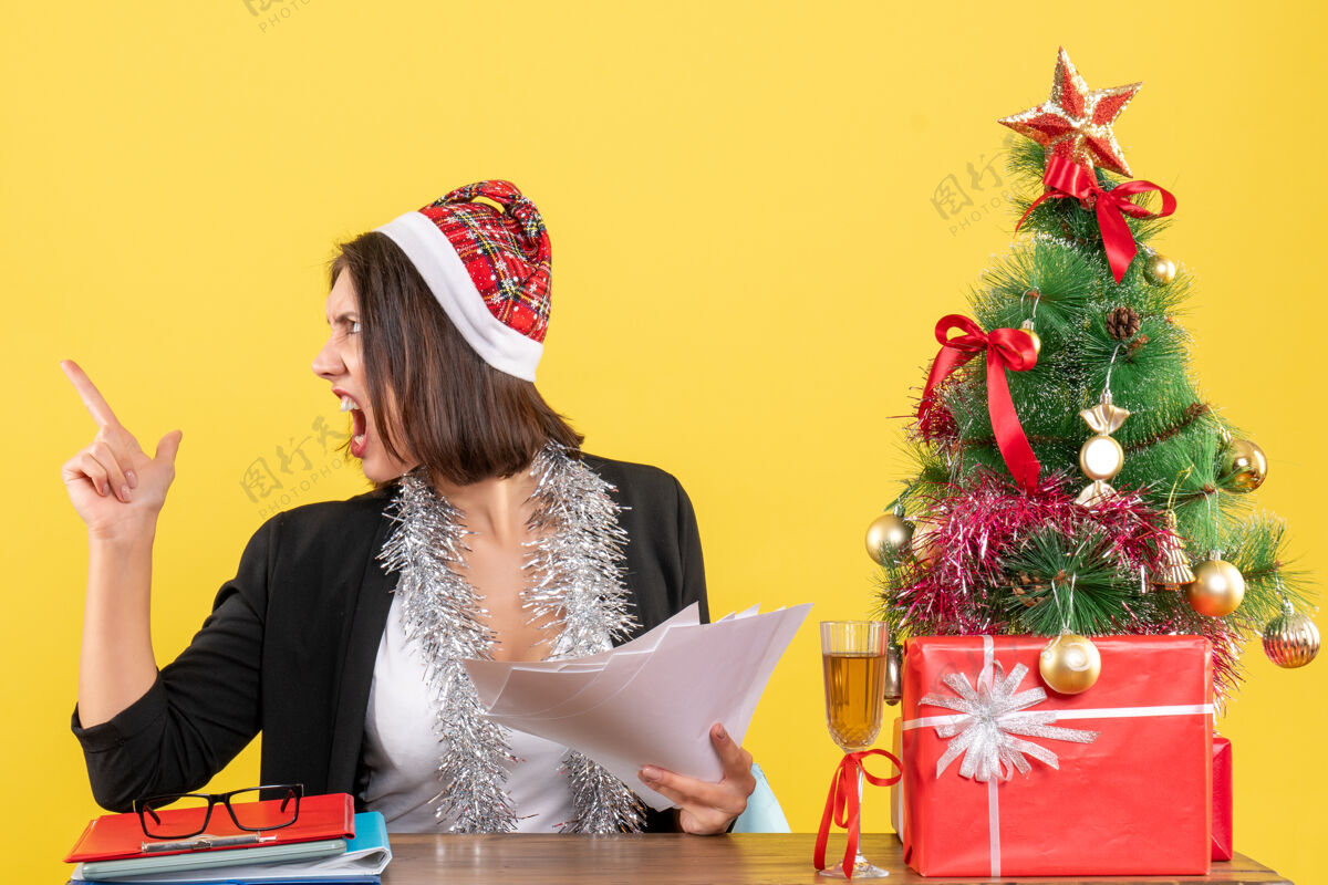 文件情绪紧张的商务女士穿着西装 戴着圣诞老人帽和新年装饰品 手里拿着文件 坐在一张桌子旁 桌子上放着圣诞树年份树帽子