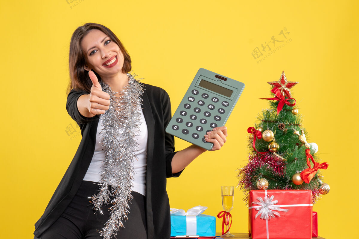 计算器圣诞气氛 微笑的美丽女士站在办公室 指着计算器在黄色的办公室做ok手势漂亮指点肖像