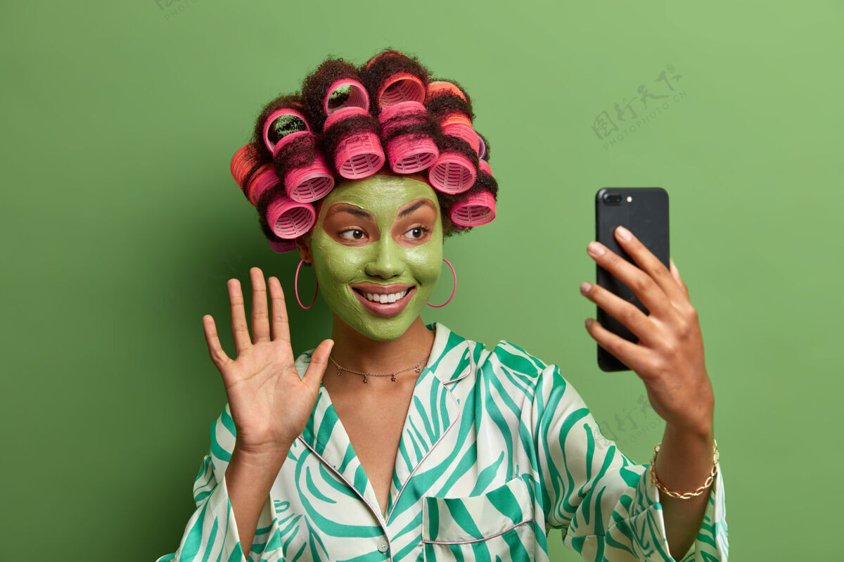 治疗快乐的女性模特 戴着绿色的面膜 挥动手掌 向朋友问好 通过现代智能手机召开视频会议 佩戴发卷 做完美发型 穿休闲装治疗室内年轻