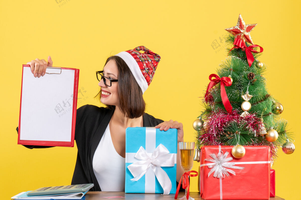 黄色迷人的女士穿着西装 戴着圣诞老人的帽子和眼镜 手里拿着文件指向办公室里的礼物帽子拿着文件