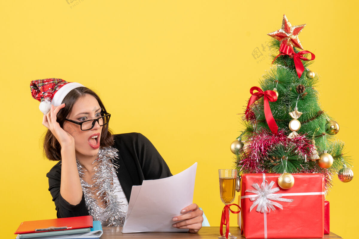 坐穿着西装 戴着圣诞老人帽和新年装饰品的商务女士 情绪激动地坐在办公室的一张桌子旁 桌上放着圣诞树肖像感情美丽