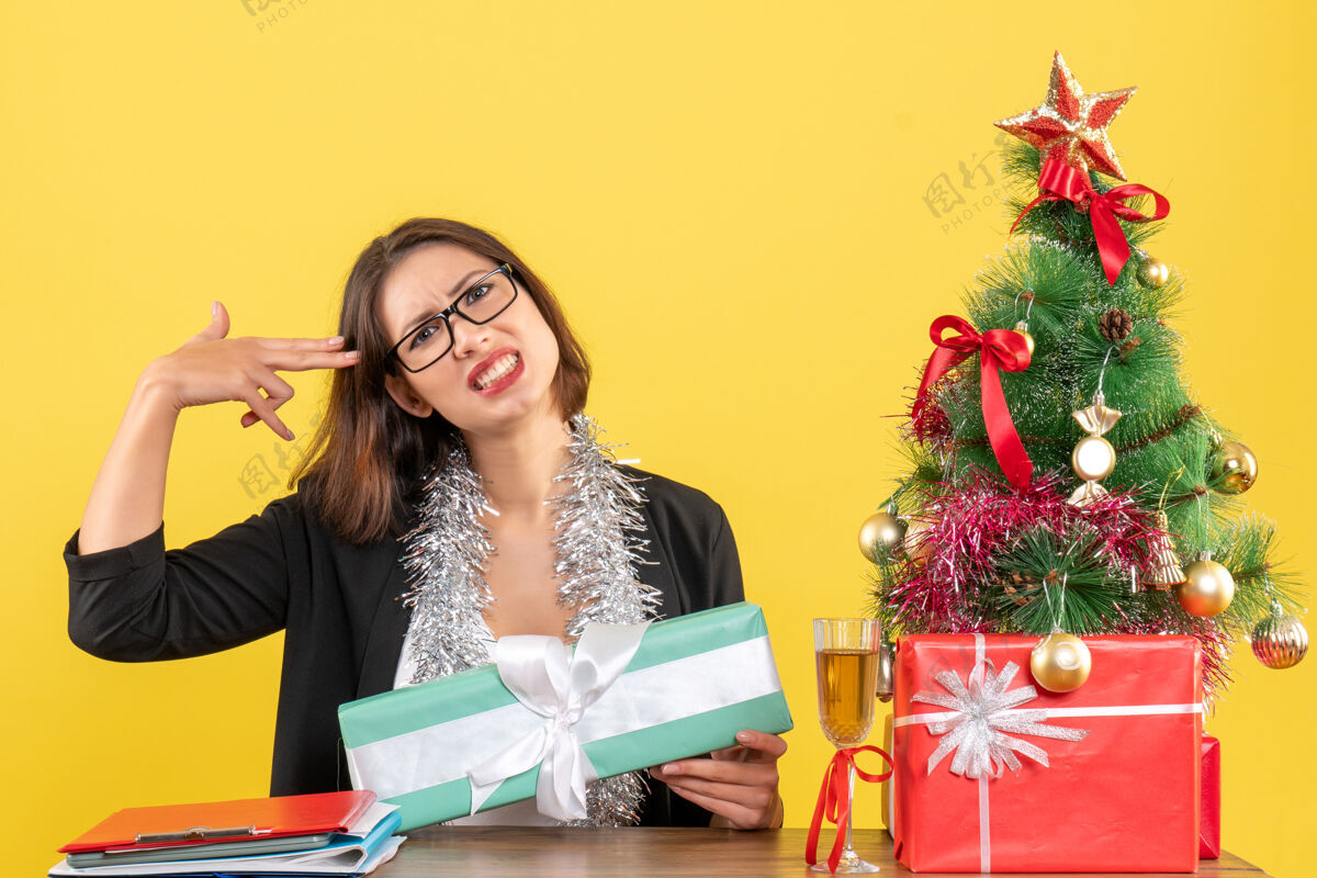 时尚一位穿着西装 戴着眼镜的商务女士 她拿着礼物坐在办公室的一张桌子旁 上面有一棵圣诞树成人漂亮困惑