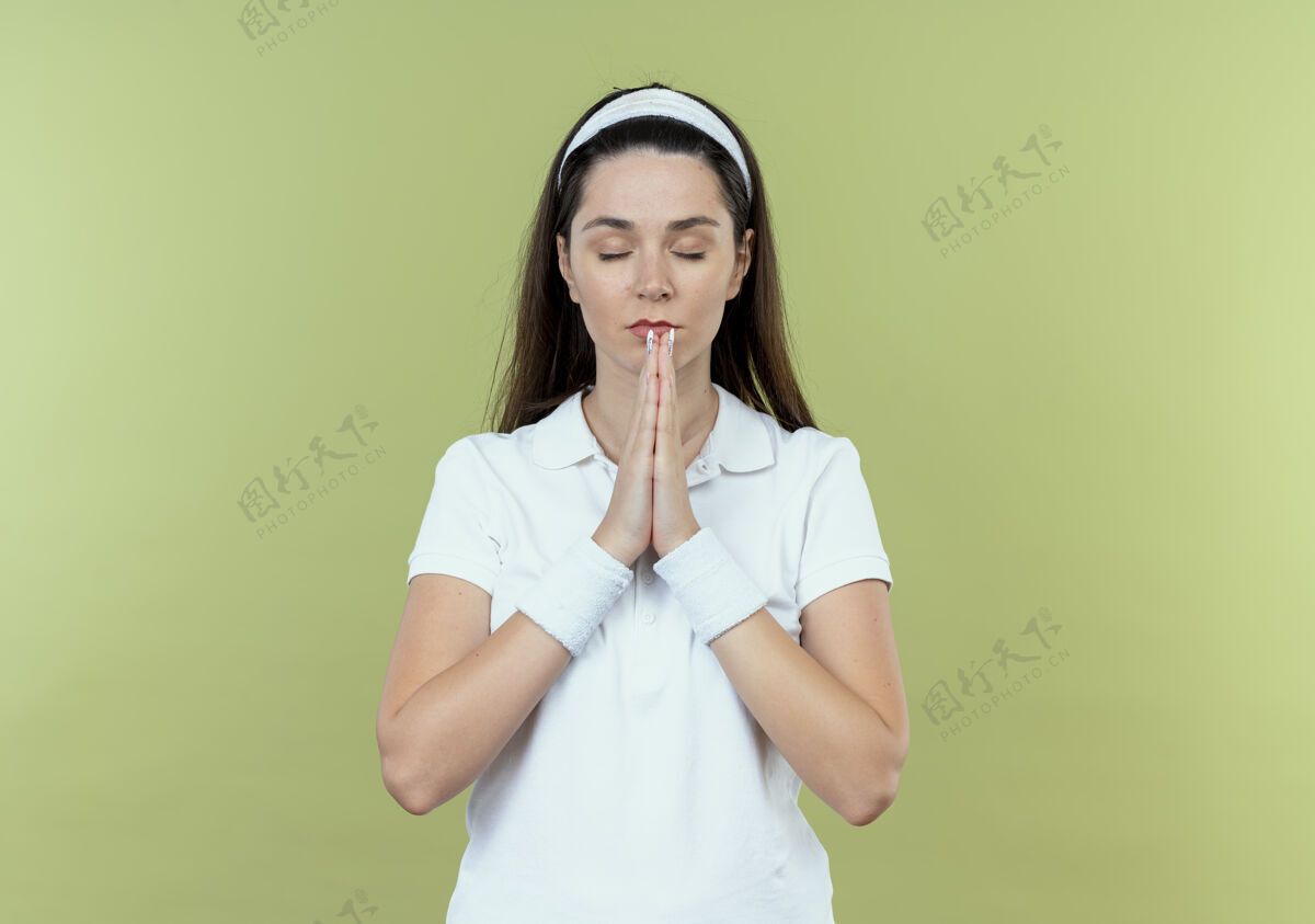 像戴着头巾的年轻健身女士双手合十 闭着眼睛站在灯光背景下祈祷健身手在一起