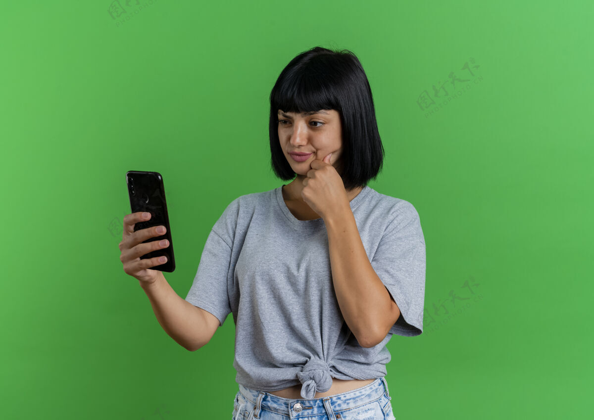 下巴困惑的年轻黑发高加索女孩把拳头放在下巴上看着绿色背景上的手机和复制空间空间拳头绿色