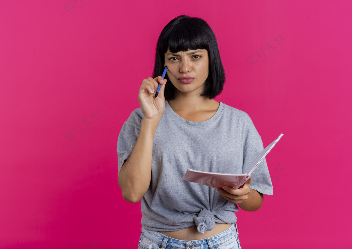 女孩体贴的年轻黑发白种女孩把笔放在脸上 拿着笔记本 看着相机隔离在粉红色的背景和复印空间持有空间钢笔