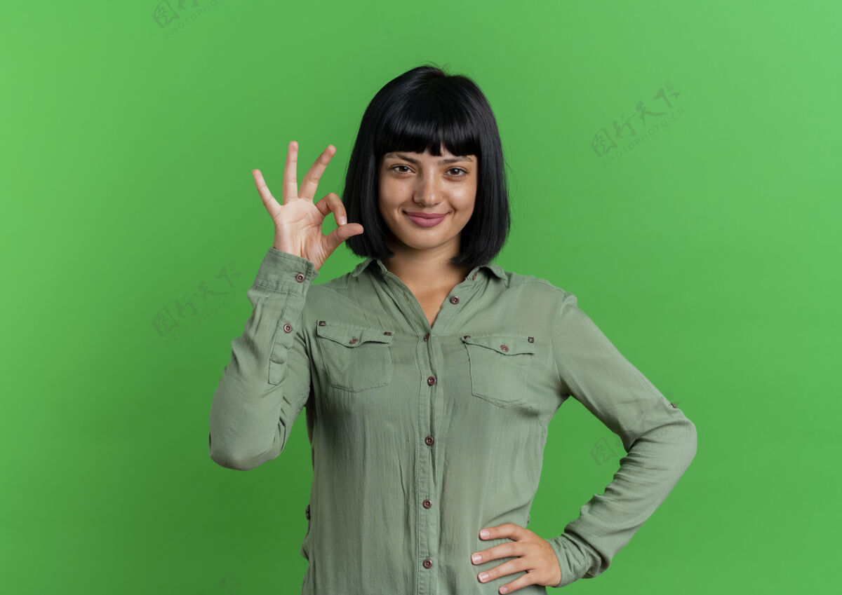 黑发高兴的年轻黑发白人女孩把手放在腰上做手势ok手牌隔离在绿色背景和复制空间标志空格女孩