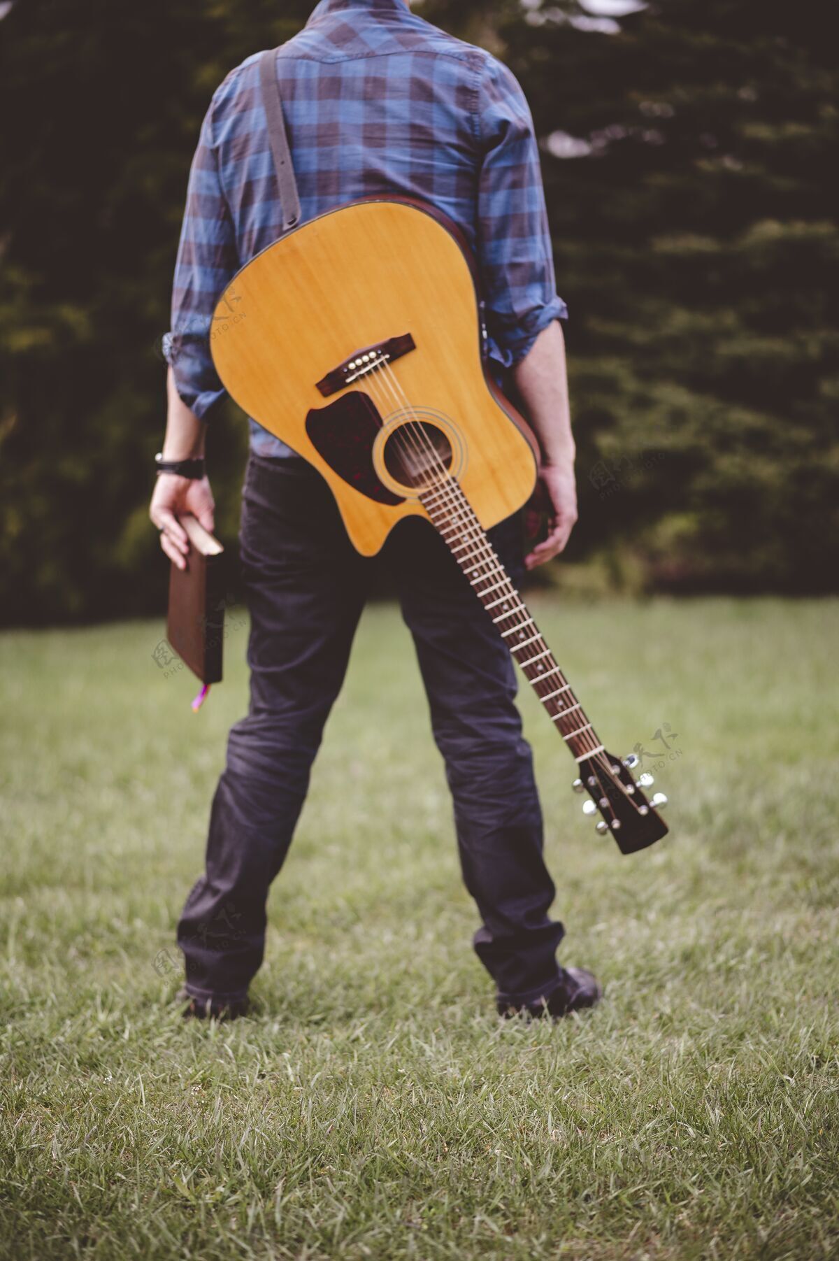 年轻一个手拿吉他和书站在草地上的男人的垂直镜头焦点弦乐宗教