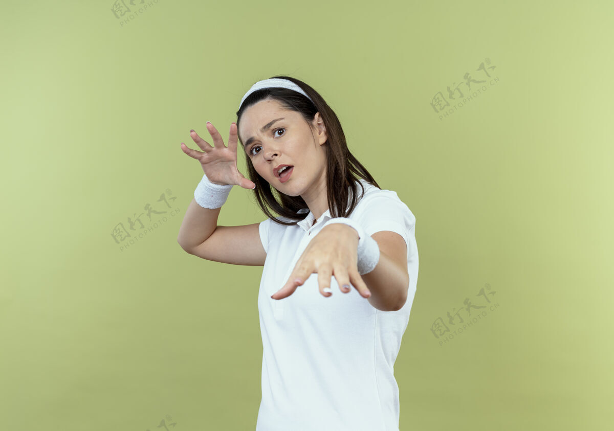 年轻戴着头巾的年轻健身女做着防御手势 双手看着摄像机 恐惧的表情站在灯光背景下站立手势相机