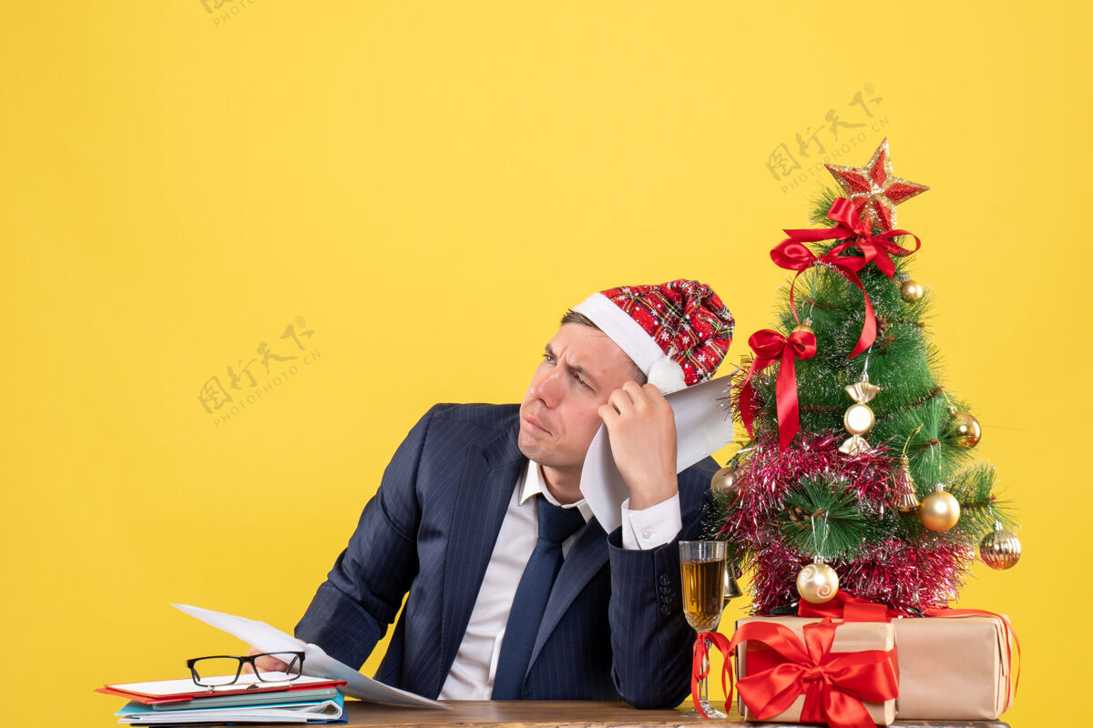 等等前视图激动的男人想的东西坐在圣诞树附近的桌子上 黄色背景上的礼物笔记本电脑商务办公室