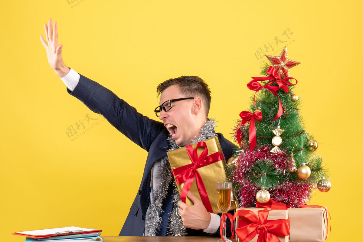 人前视图愤怒的男人张开手坐在圣诞树旁的桌子上 呈现黄色背景前面漂亮打开
