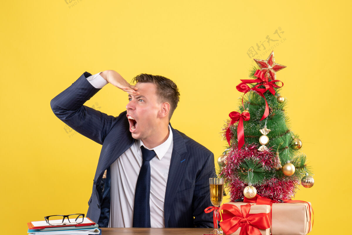 行政前视图愤怒的男人坐在圣诞树旁的桌子旁 在黄色背景下呈现新郎附近坐着