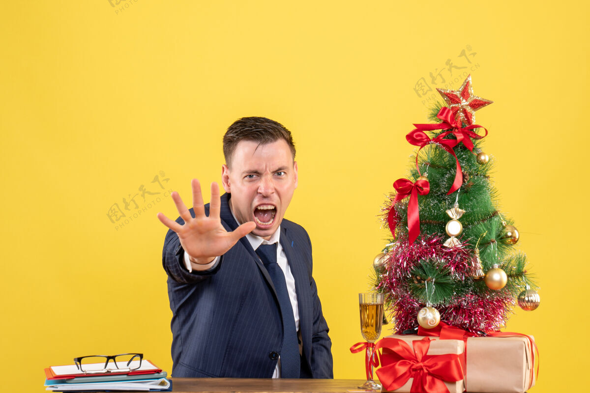停止前视图愤怒的男人停下手坐在圣诞树旁的桌子上 呈现黄色背景靠近手新郎