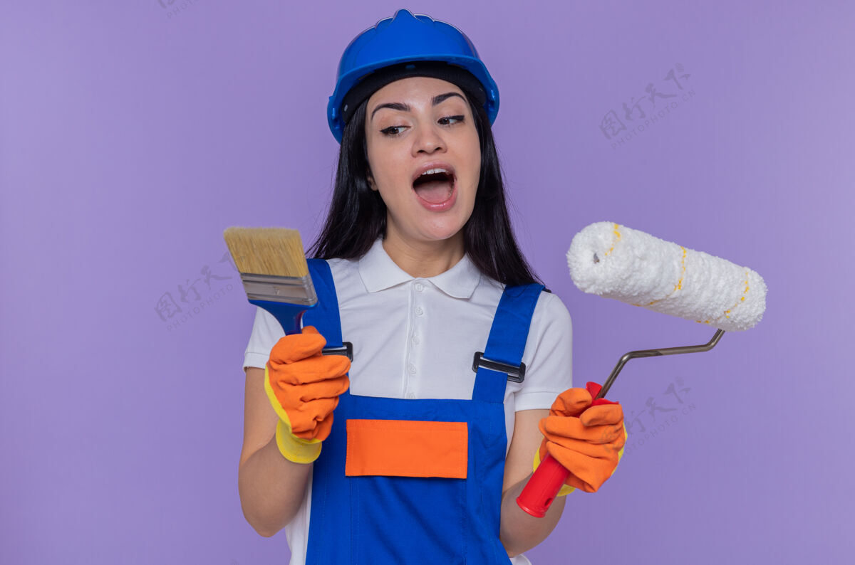 滚轴年轻的女建筑工人穿着施工制服 戴着安全帽 戴着橡胶手套 拿着油漆辊和刷子 看上去很惊讶油漆年轻人制服