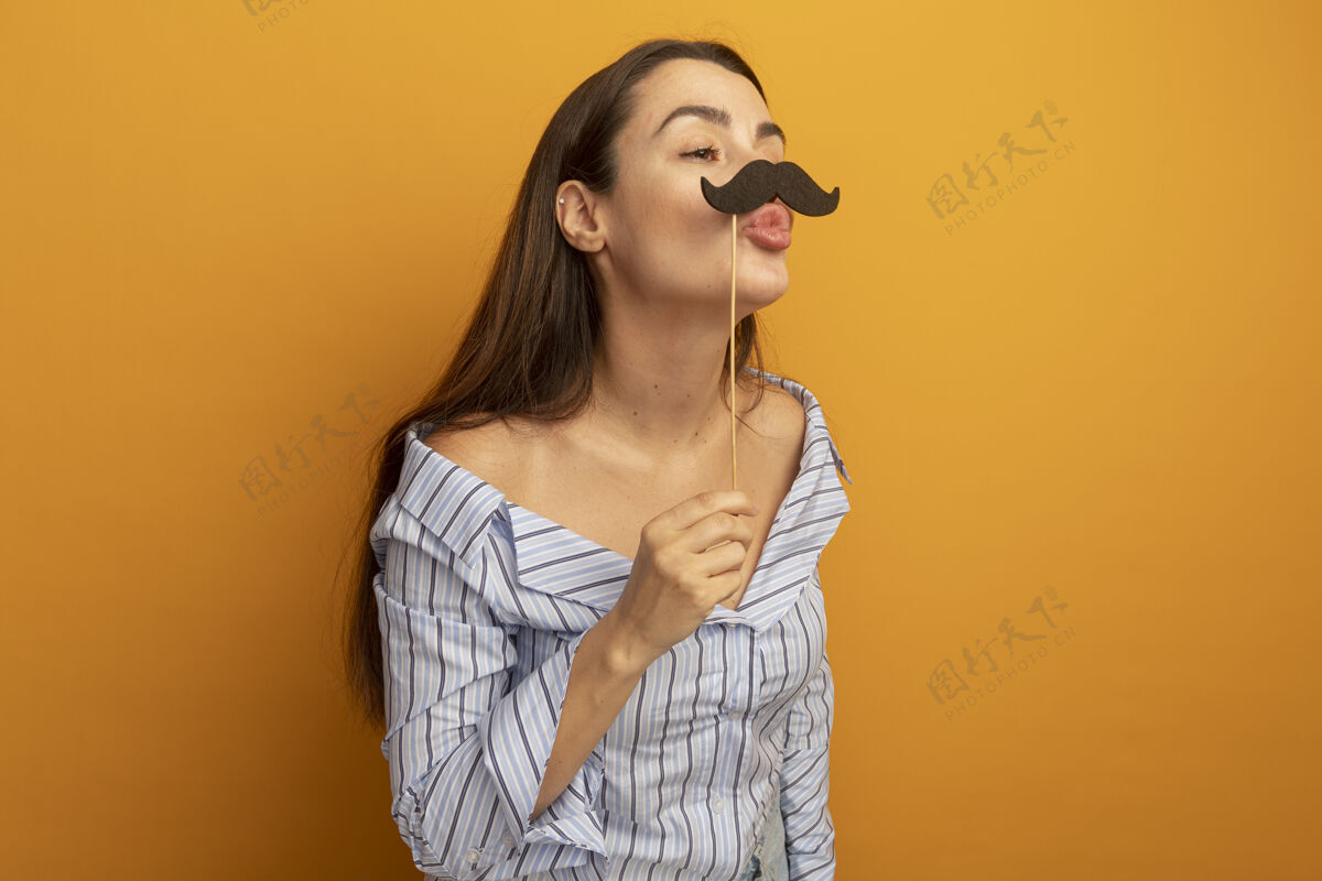 胡子高兴的美女拿着假胡子在棍子上看着橙色墙上孤立的一面壁板人表情